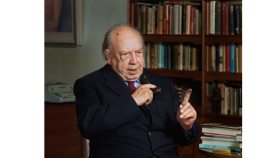 Portrait of JB Preistley - an older white male dressed in a navy blue suit, light blue shirt and red tie, sitting in a chair with bookshelves in the background diaganol to the camera