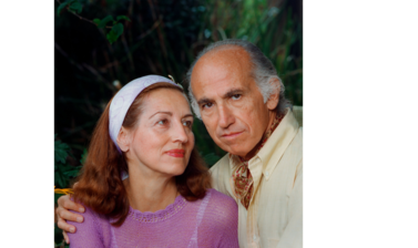 Portrait of Jonas Salk and Françoise Gilot - an older white male balding with white hair, dressed in a cream shirt and jumper with a patterned neckscarf, hs arm is round a young red haired woman in a pink jumper and white headband.