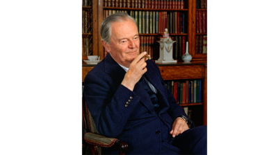 Portrait of Kenneth Clark - an older white male with grey hair, dressed in a navy blue suit, blue stripey shirt and navy tie, sitting in a wooden chair looking diagonal to the camera with bookshelves in the background and a cigar in his hand