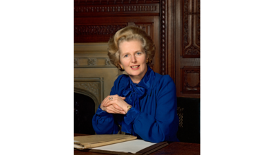 Portrait of Margaret Thatcher - a middle aged white woman with light brown hair, dressed in a bright blue silk blouse, sitting at a desk in a wooden panelled room with hands clasped together looking into the camera