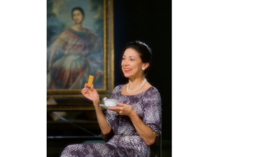 Portrait of Margot Fonteyn - a middle aged white woman with dark hair up in a bun, wearing a purple and white flower patterned dress, sitting in a chair with a cup of tea and biscuit in hand, sat in front of a large oil painting