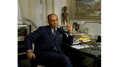 Portrait of Ralph Ellison - an older black male wearing a navy suit, blue shirt and navy striped tie, sitting next to a desk in an office diagonal to the camera
