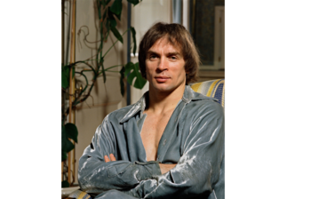 Portrait of Rudolf Nureyev - a young white man with light brown hair sat on a sofa with a light blue velvet shirt on