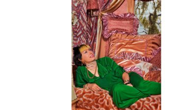 Portrait of Zandra Rhodes - a middle aged white woman with dark hair and a blonde fringe, wearing a green jumpsuit, laying on a sofa covered with peach blankets and cushions, looking upwards