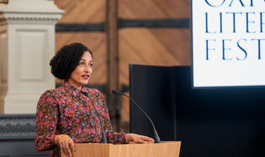 Zadie Smith speaks from a podium in front of a blue background, beside a screen