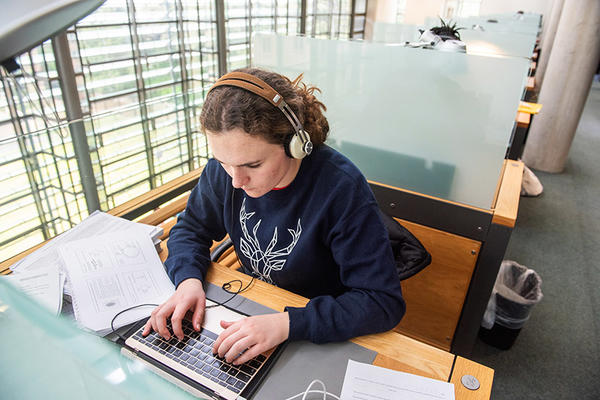 A woman wearing headphones works at a laptop, with papers on the desk around her
