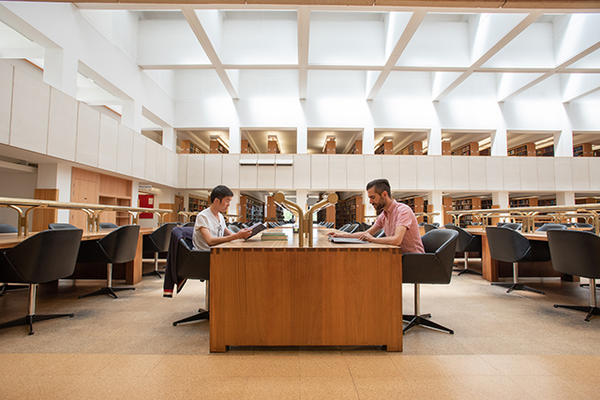 A double height reading room that is light and airy - there is a long wooden desk and two students sit either side working