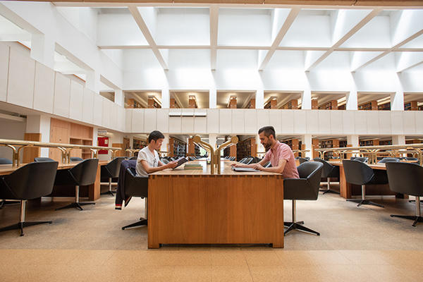 A double height reading room that is light and airy - there is a long wooden desk and two students sit either side working