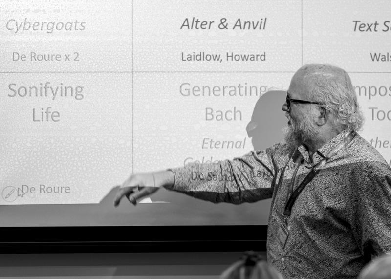 A black and white photograph of an older man with a beard and glasses standing in front of a projection of a slide