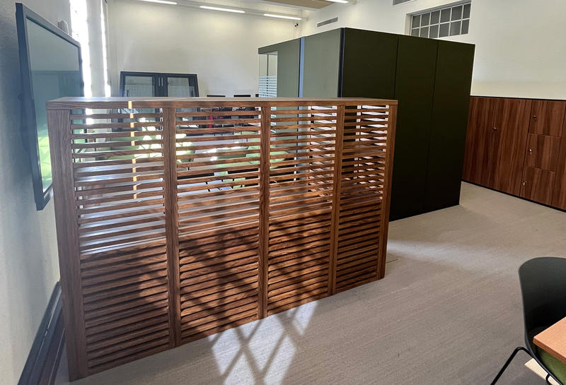 A view of a slatted wooden screen with a table and two sofas behind it; to the right is a large green pod booth for a private workspace 