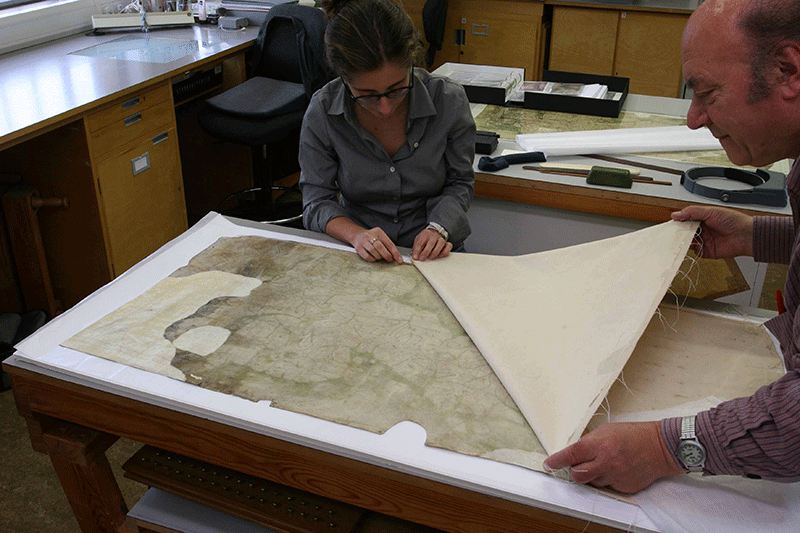 The Gough Map is flat on a table - two people sit next to it and peel off a textile back lining