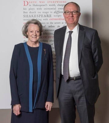 A photograph of a woman with a long bob in a navy suit and a tall man with glasses in a grey suit