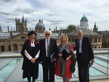 Dame Hilary Mantel in a red and grey academic gown standing on rooftop with Oxford skyline behind. She is accompanied by a woman in full academic dress and two men in dark suits.