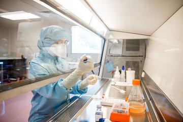 A scientist in full PPE clothing - they are holding a test tube in pipette in each hand behind a screen. Various flasks and containers are behind the screen