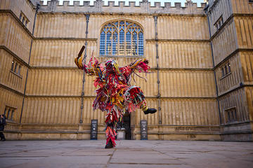 A Black man wearing a colourful costume made from strips of material dances in front of the entrance of the Bodleian Old Library