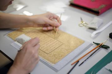 A detail of a person's hands over a manuscript document. They hold a wooden tool in each hand and delicately work on the document
