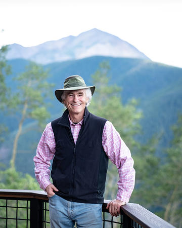 A man wearing a dark jacket and purple shirt stands on a balcony in front of a mountain range