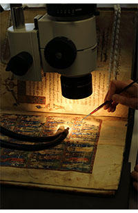 A microscope is seen above an illuminated manuscript - a light shines down on the illumination and a paint brush is positioned close to the surface