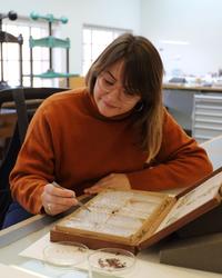 A woman holds what looks like a pair of tweezers and moves them towards a historic book - in front of the book are two petri dishes