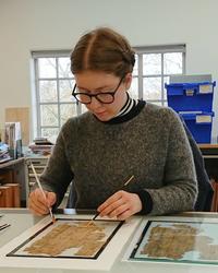 A woman sits at a desk - she has a paint brush in each hand and is working on a fragile manuscript that is broken into many pieces