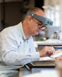 Andrew Honey A side profile of a man - he has microscope glasses on his head - he holds a brush in his hand and works on a historic book