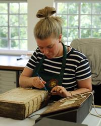A woman sits at a desk wearing an apron. In front of her is a large book with its cover off - she is using an implement to conserve the spine of the book