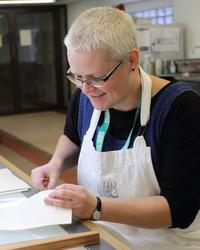 A woman wears a white apron, in her hands she is holding a piece of paper that she is sewing