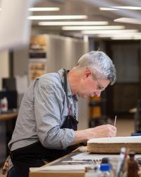 Simon Haigh A side profile of a man - he is sitting down at a desk looking down, a book is open in front of him and he works on it with a small implement